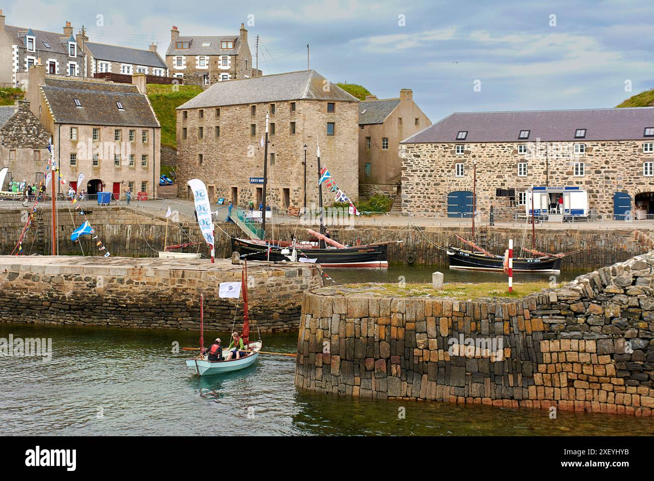 Portsoy Boat Festival ein kleines Ruderboot mit braunen Segeln und zwei Seeleuten, die den alten Hafen betreten Stockfoto