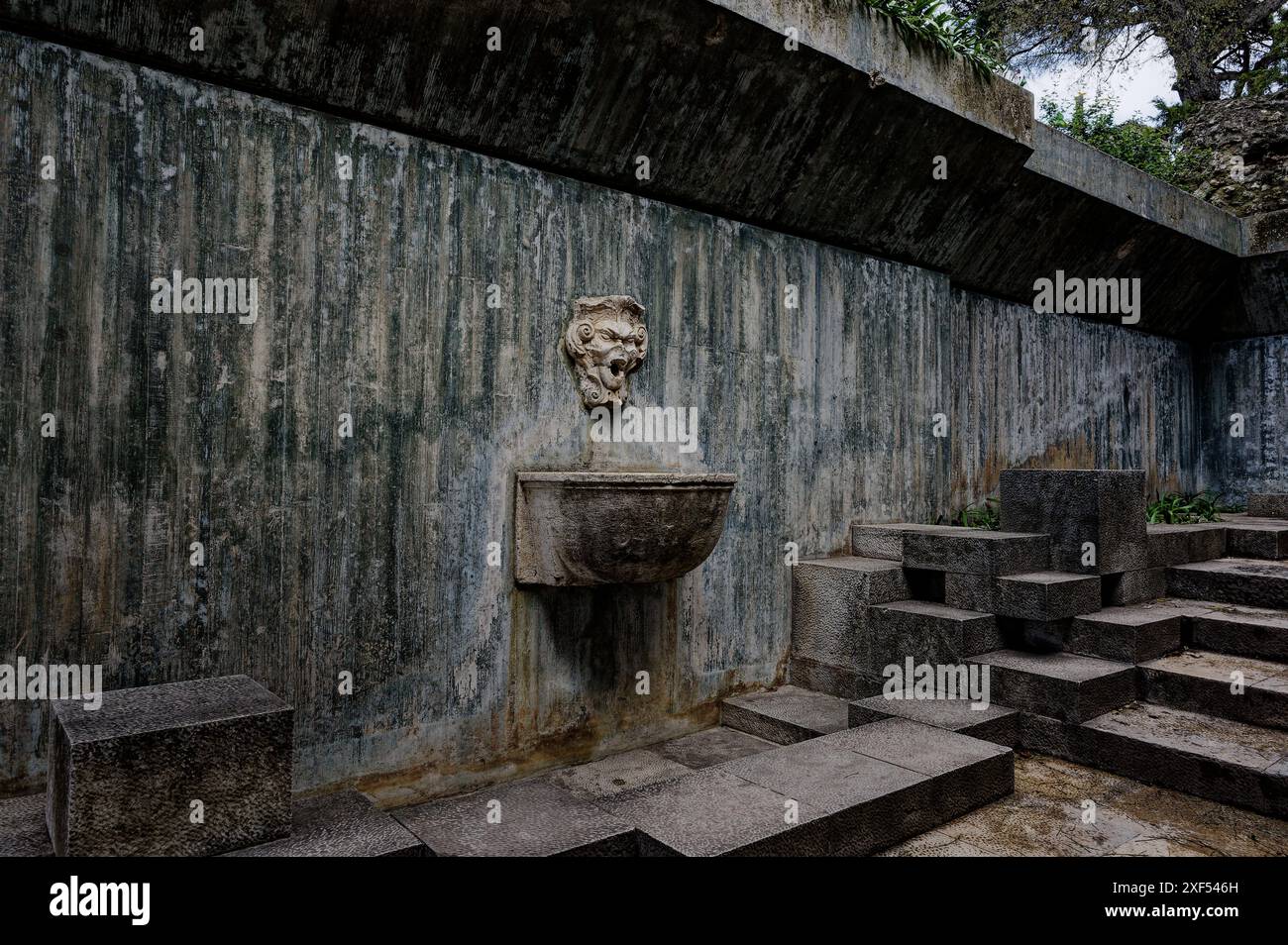 Verwitterter Steinbrunnen mit grotesker Fassade im historischen Castelo de São Jorge, Lissabon, vor alten Mauern und modernen Steintreppen Stockfoto