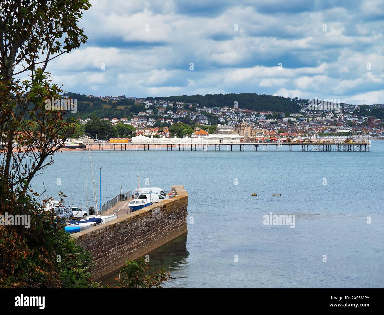 Ein Blick auf den Paignton Pier, die Häuser am Meer und am Hügel, ein Teil der Hafeneingangsmauer befindet sich im Vordergrund, während der Himmel mit Cumulus cl beladen ist Stockfoto