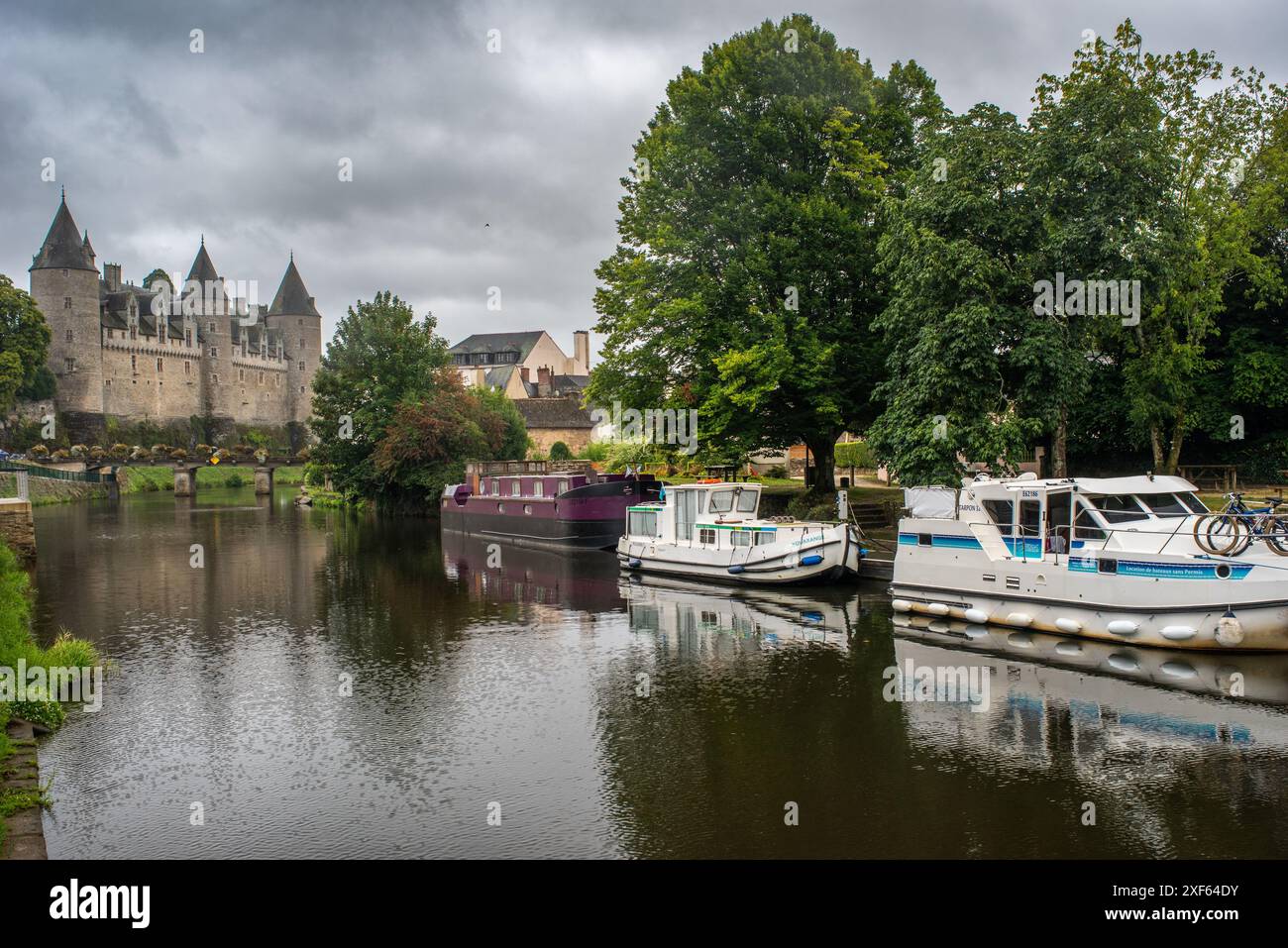 Malerische Aussicht auf Boote, die auf dem ruhigen Oust River liegen, mit dem historischen Schloss Josselin im Hintergrund, das sich in Josselin, Bretagne, Frankreich befindet. Stockfoto