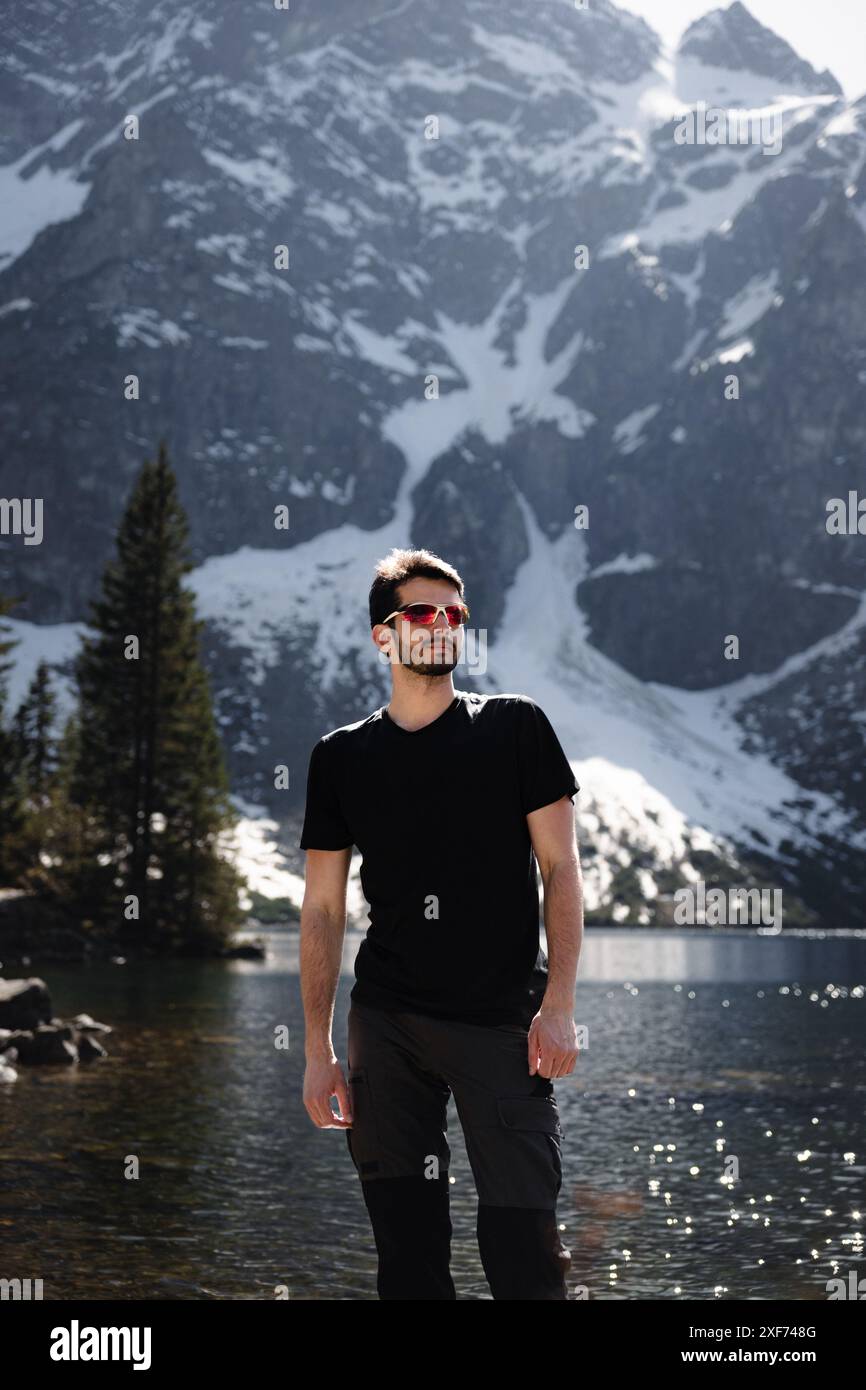 Mann in schwarzem Hemd und Sonnenbrille am See mit Bergkulisse Stockfoto