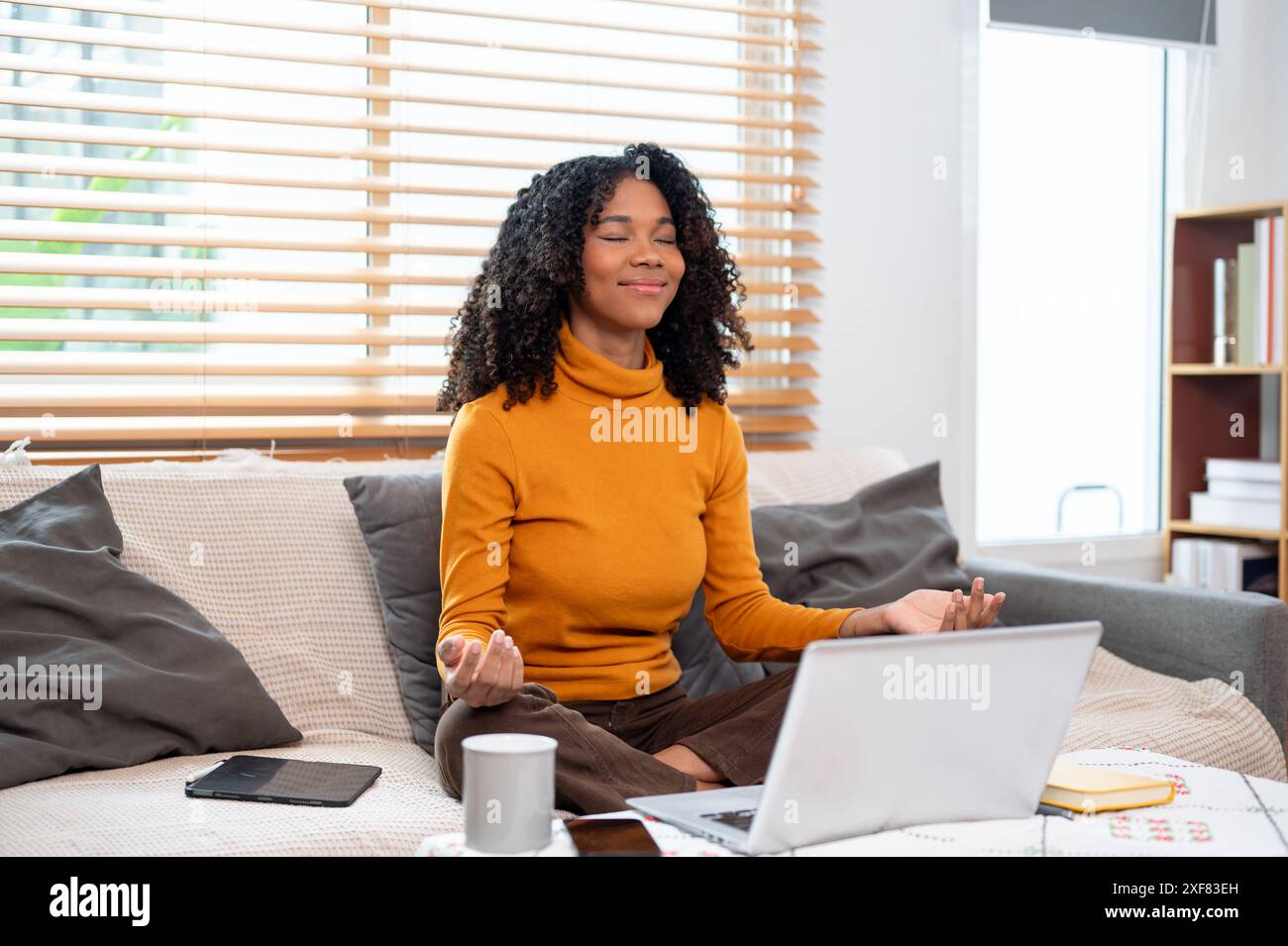 Eine ruhige, positive schwarze Frau meditiert auf einer Couch im Wohnzimmer, bevor sie mit der Arbeit beginnt. Achtsamkeit, positive Einstellung, Entspannung, Stresssituation Stockfoto