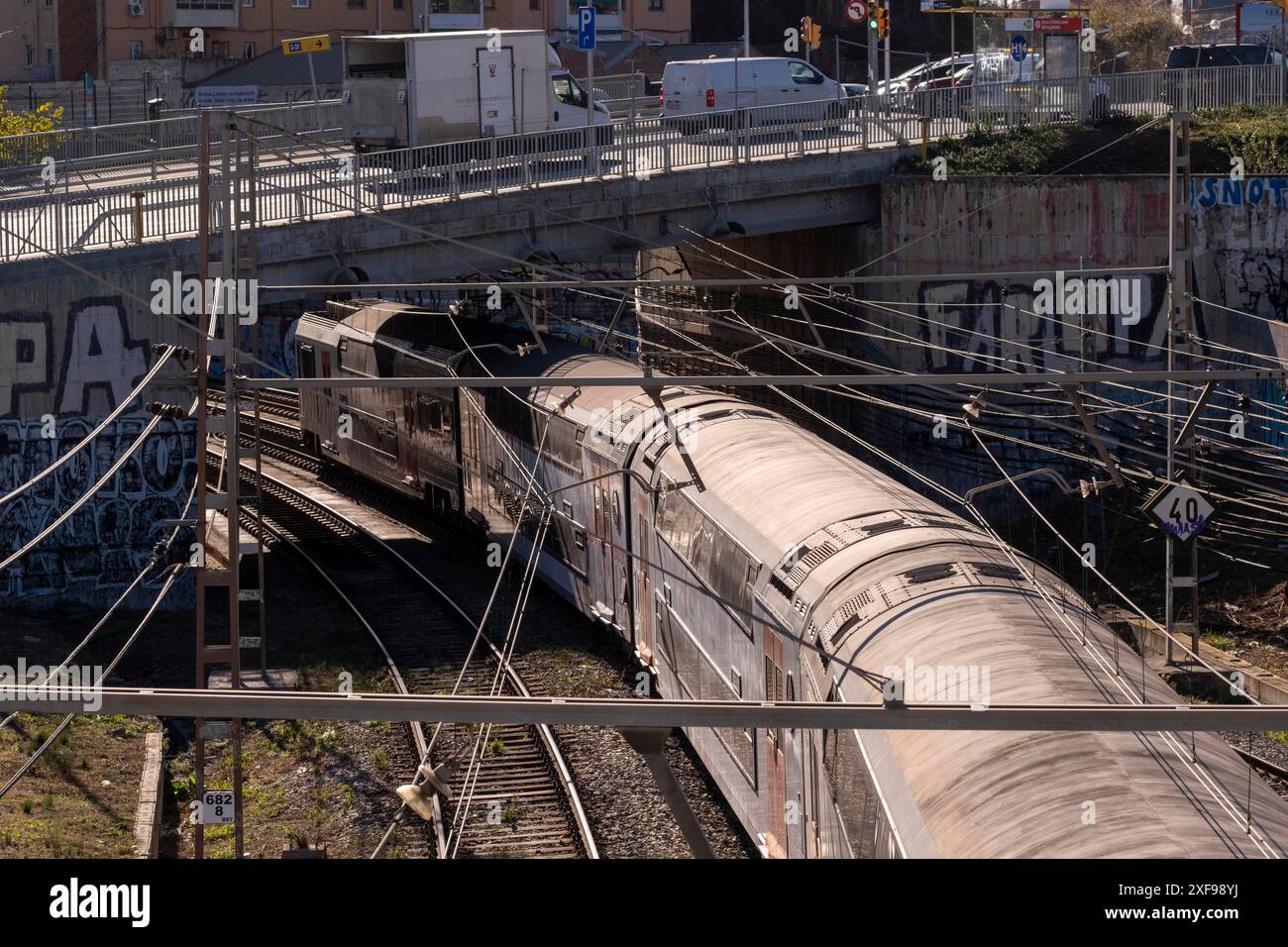 Zug, der unter einer mit Graffiti bedeckten Brücke auf Eisenbahngleisen in einer städtischen Umgebung vorbeifährt, Poblenou, Barcelona, Katalonien, Spanien Stockfoto