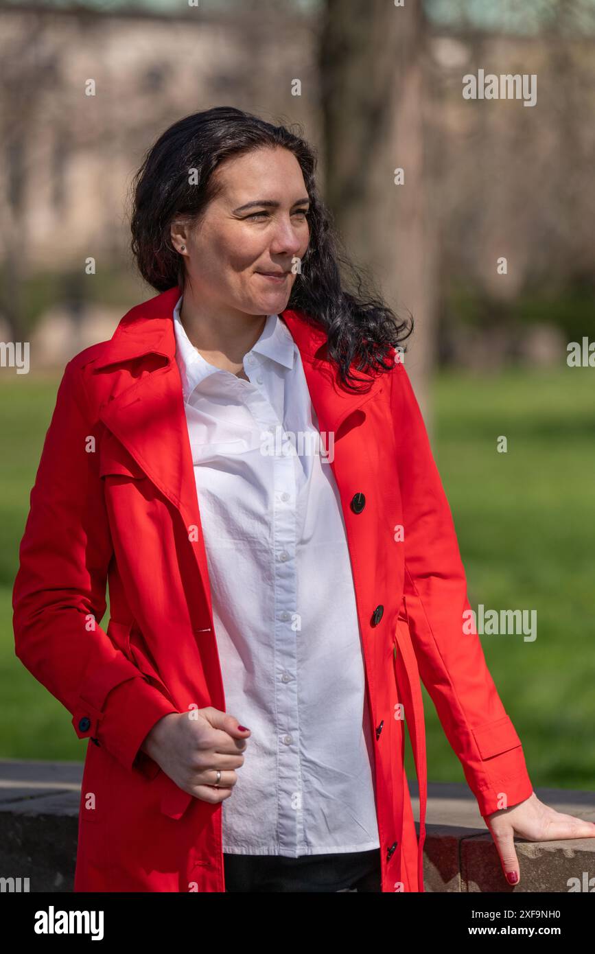 Porträt einer Frau mit dunklem Haar in rotem Regenmantel und Geschäftsbluse. Er steht unter freiem Himmel und blickt nachdenklich in die Ferne. Stockfoto