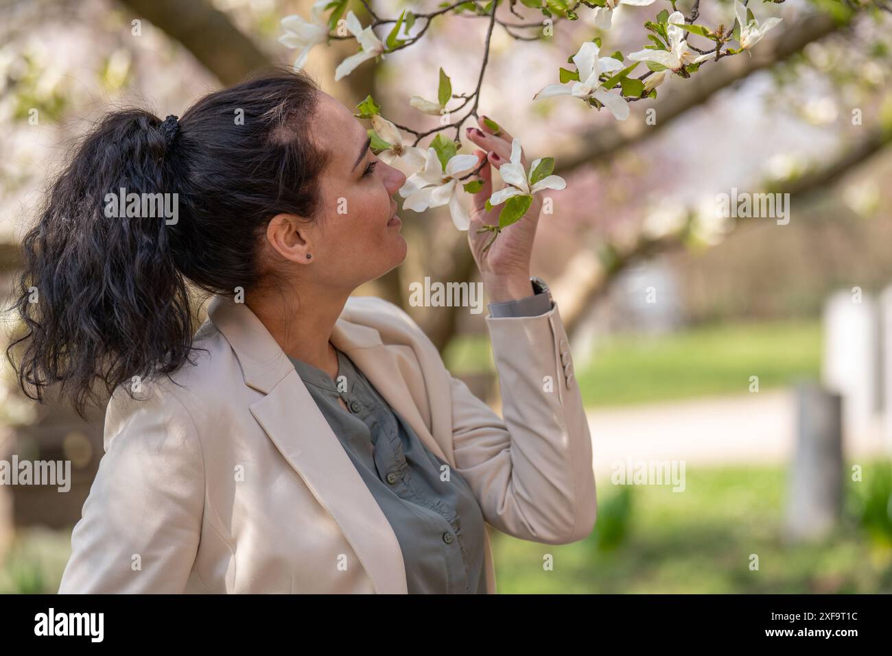 Romantisches Bild einer stilvollen Frau in einer leichten Jacke. Positive Frühlingsstimmung. Ein süßes Mädchen hält sanft einen weißen Sakura-Zweig und schaut sich die Blumen an Stockfoto