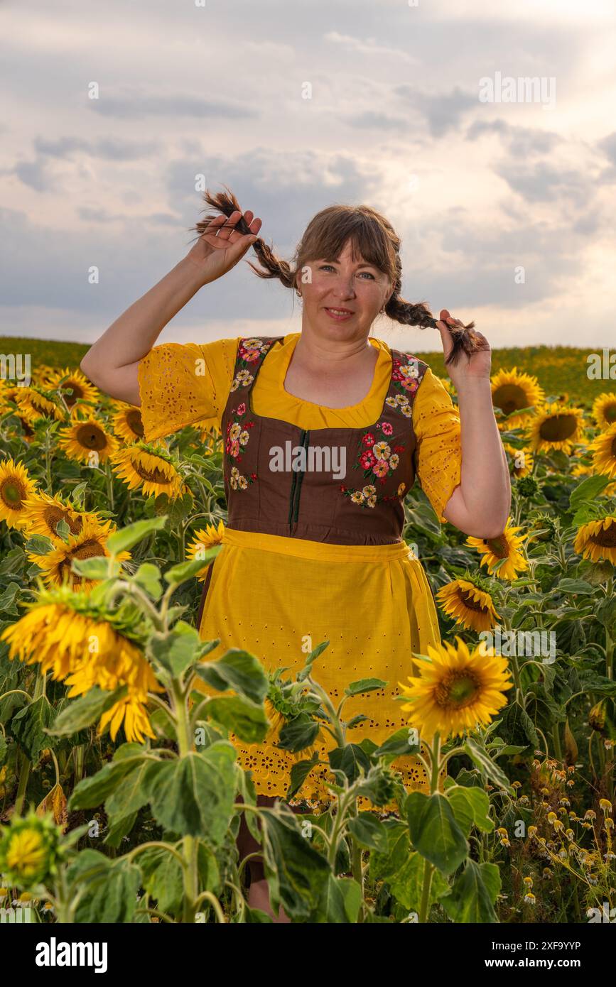 Eine Frau steht mitten auf einem Feld mit vielen gelben Sonnenblumen. Geflochtene Haare, gekleidet in deutscher Nationalkleidung, gelbe Bluse, brauner Sundr Stockfoto