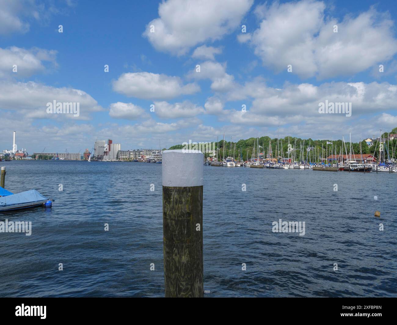 Hafen mit Booten und Holzpfosten, im Hintergrund ein Industriegebiet und bewaldete Hügel unter einem blauen Himmel mit Wolken, Stein, schleswig. deutschland Stockfoto