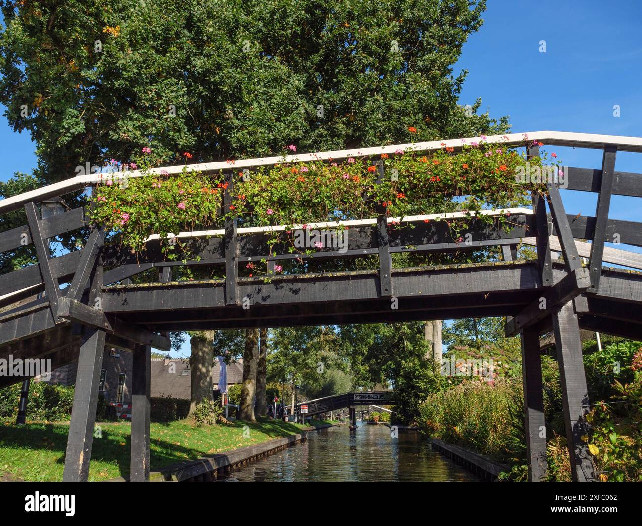 Holzbrücke mit hängenden Blumen über einem ruhigen Kanal, umgeben von grünem Laub und einem hellblauen Himmel, giethoorn, niederlande Stockfoto