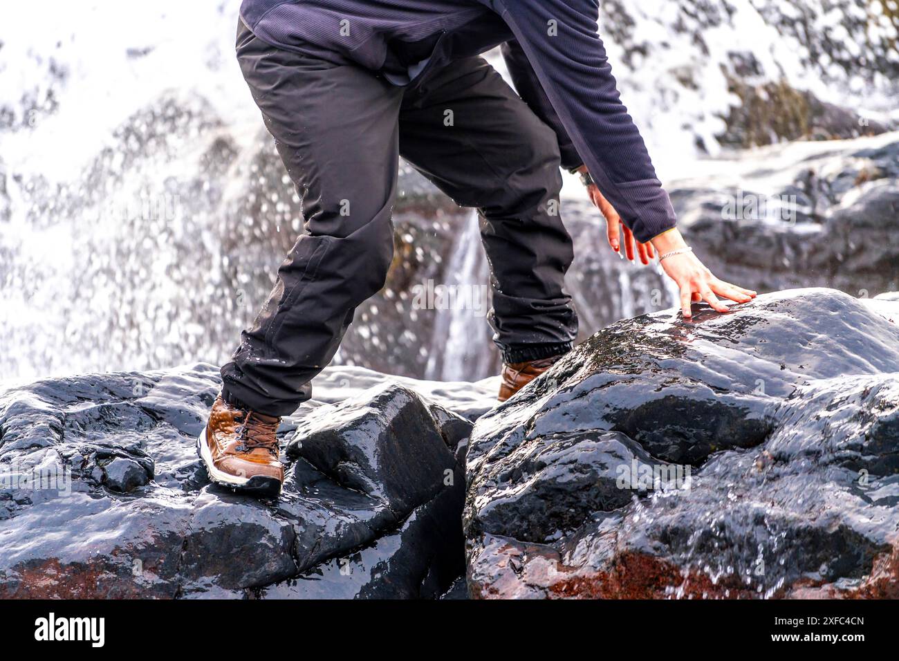 Ein Mann steht auf einem Felsen mit der Hand auf einem nassen Felsen. Der Mann trägt eine schwarze Jacke und eine schwarze Hose. Abenteuer- und Entdeckungskonzept Stockfoto