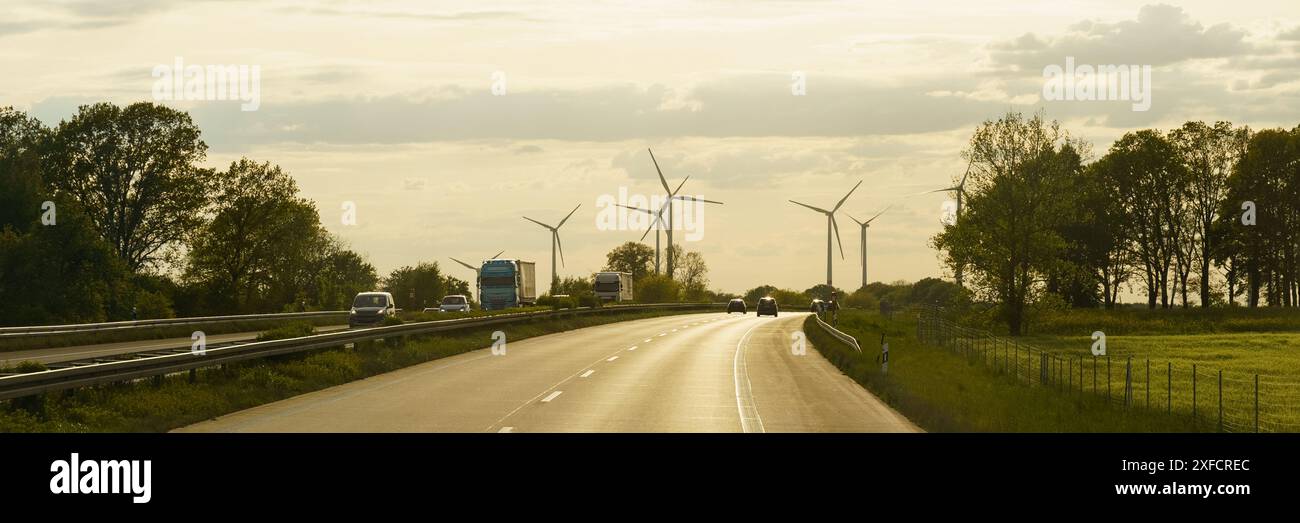 Eine asphaltierte Straße schlängelt sich durch eine ländliche Landschaft mit Bäumen an den Seiten und einer Linie von Windrädern in der Ferne vor einem klaren Sonnenuntergangshimmel. Stockfoto