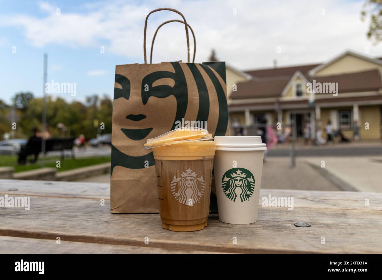 Zwei Starbucks-Drinks vor der Papiertüte - Holztisch im Freien - mit Karamell-Nieseln vereist - verschwommenes Gebäude und Baumhintergrund. Aufgenommen in Toront Stockfoto