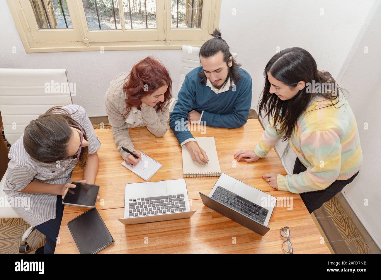 Eine Gruppe von vier Kollegen nimmt an einem gemeinsamen Meeting Teil, bei dem Laptops, Notebooks und ein Tablet zur Diskussion stehen. Stockfoto