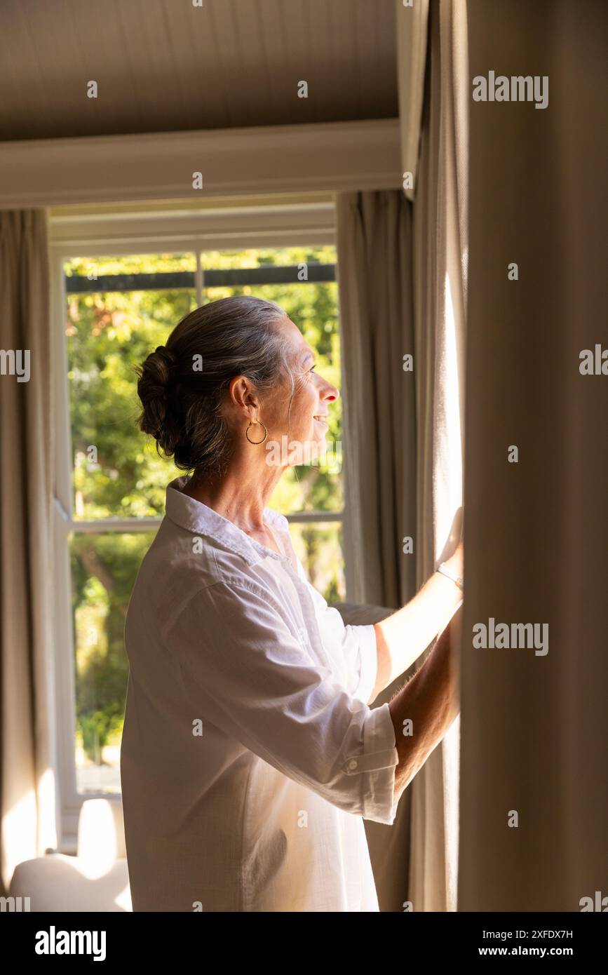 Öffnet die Vorhänge, reife Frau genießt zu Hause das Sonnenlicht und blickt nach draußen Stockfoto