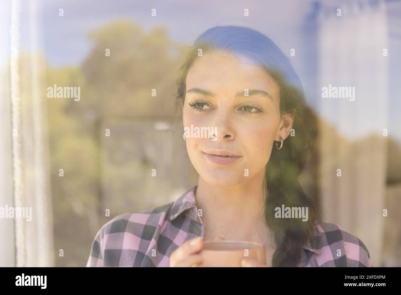 Eine junge Frau, die eine Kaffeetasse hält, aus dem Fenster blickt und einen friedlichen Moment genießt Stockfoto