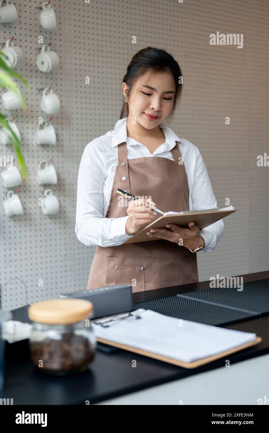 Eine attraktive junge asiatische Barista oder Kellnerin arbeitet in einem Café und schreibt Bestellungen auf die Zeitung. Service, Gastfreundschaft, kleine Unternehmen Stockfoto