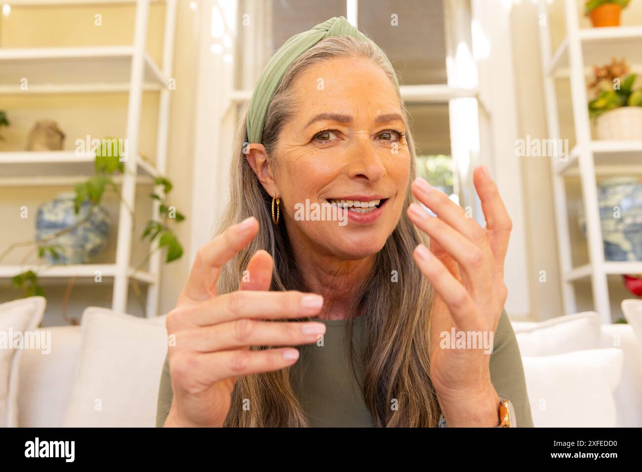 Lächelnde reife Frau, die mit Händen Gesten macht, während eines Videogesprächs auf der Couch zu Hause Stockfoto