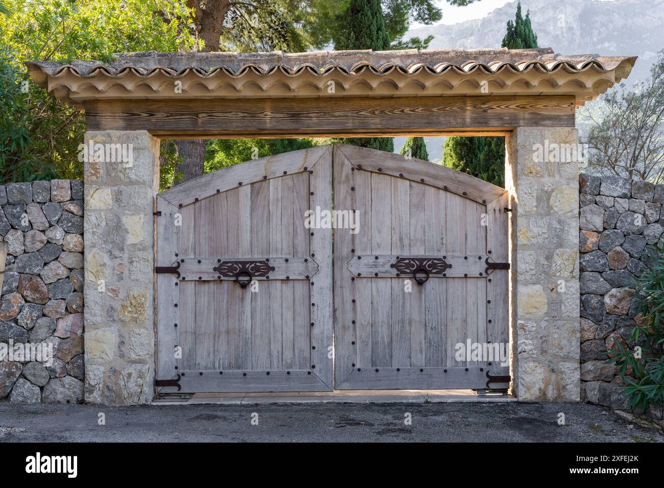 Halbrunde Holztore mit geschmiedeten Türgriffen und geschmiedeten Scharnieren. Eine Steinmauer vor dem Hintergrund majestätischer Berge. Mallorca, Spanien Stockfoto