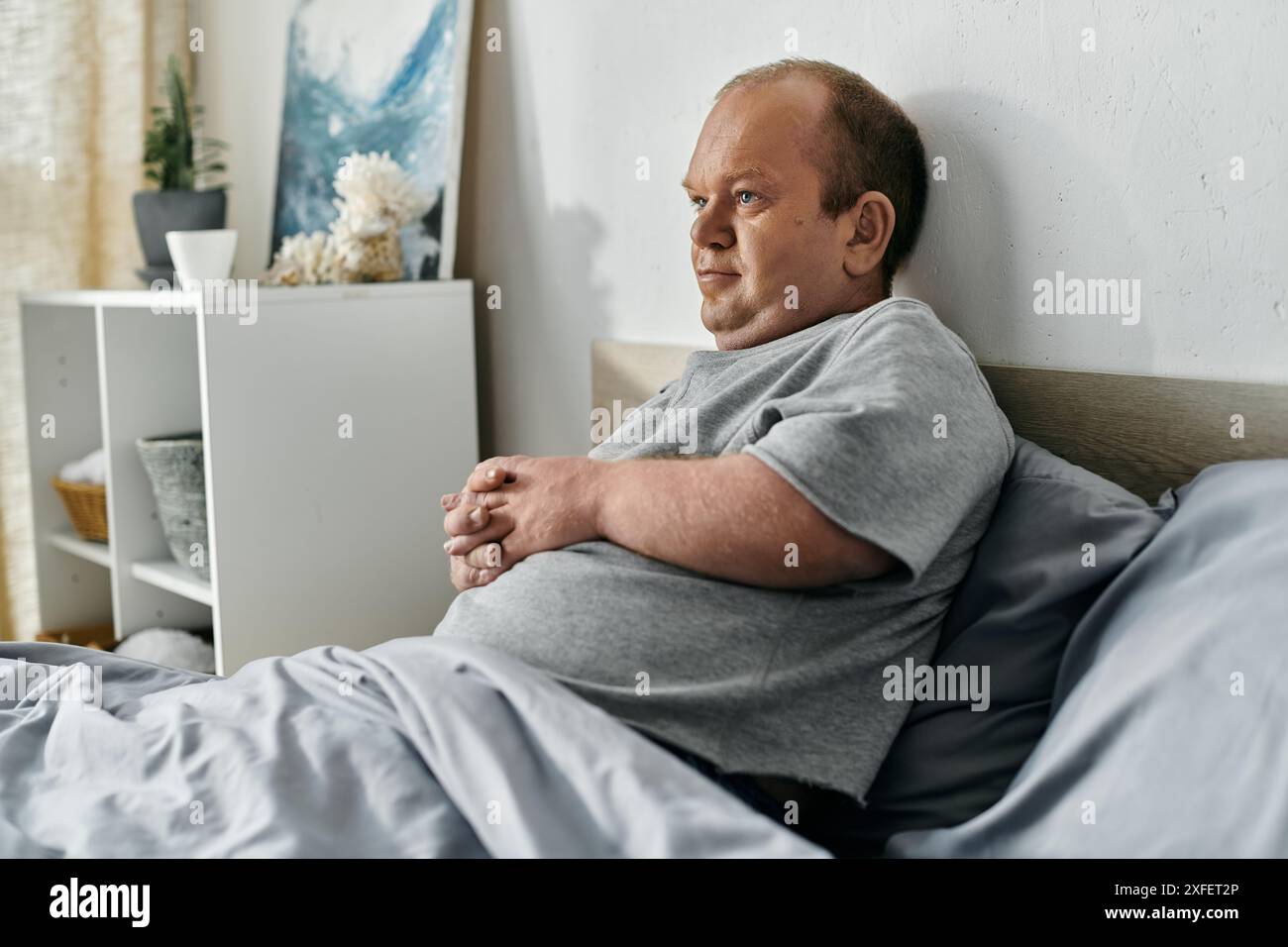 Ein Mann mit Inklusivität sitzt im Bett, verloren in Gedanken, während das Morgenlicht durch das Fenster filtert. Stockfoto