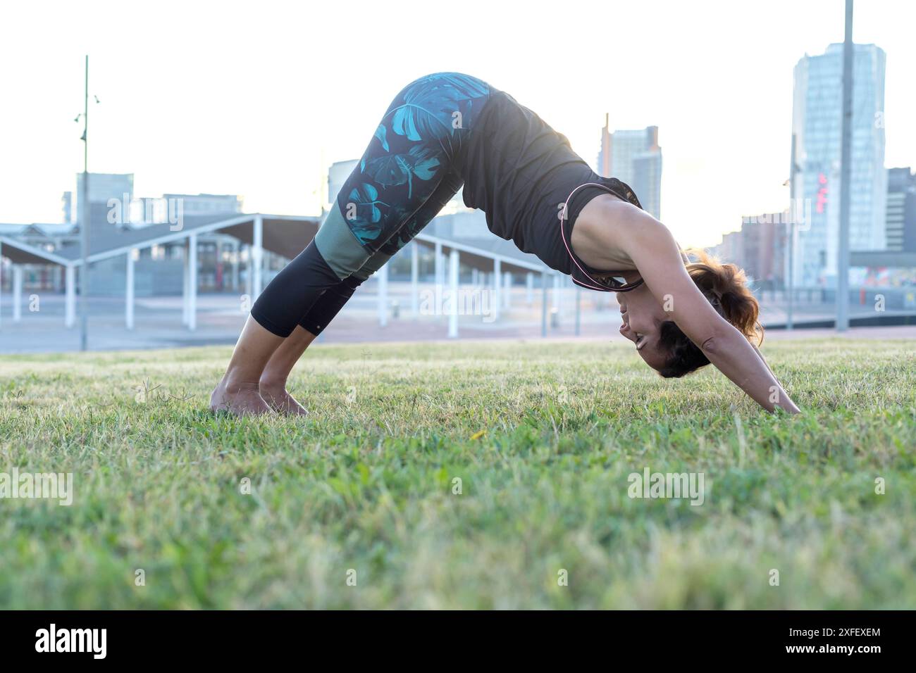 Eine Frau in athletischer Kleidung spielt auf einem grasbewachsenen Feld in einem Stadtpark die Yoga-Pose mit urbanen Gebäuden im Hintergrund. Stockfoto