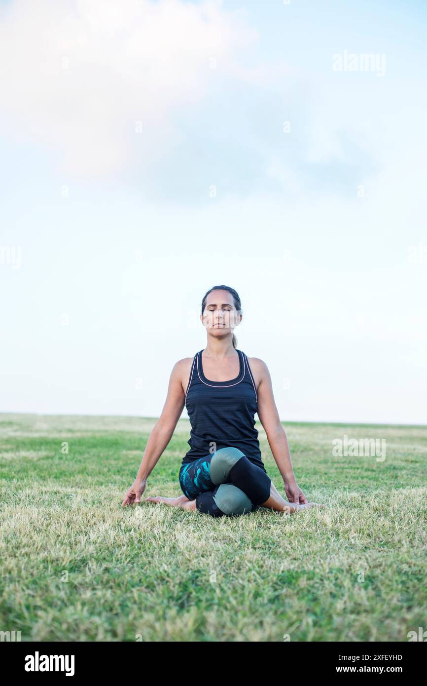 Eine Frau in sportlicher Kleidung sitzt im Kreuz auf einem grasbewachsenen Feld in meditativer Pose, mit einem ruhigen und fokussierten Ausdruck, vor einem klaren Himmel Hintergrund. Stockfoto