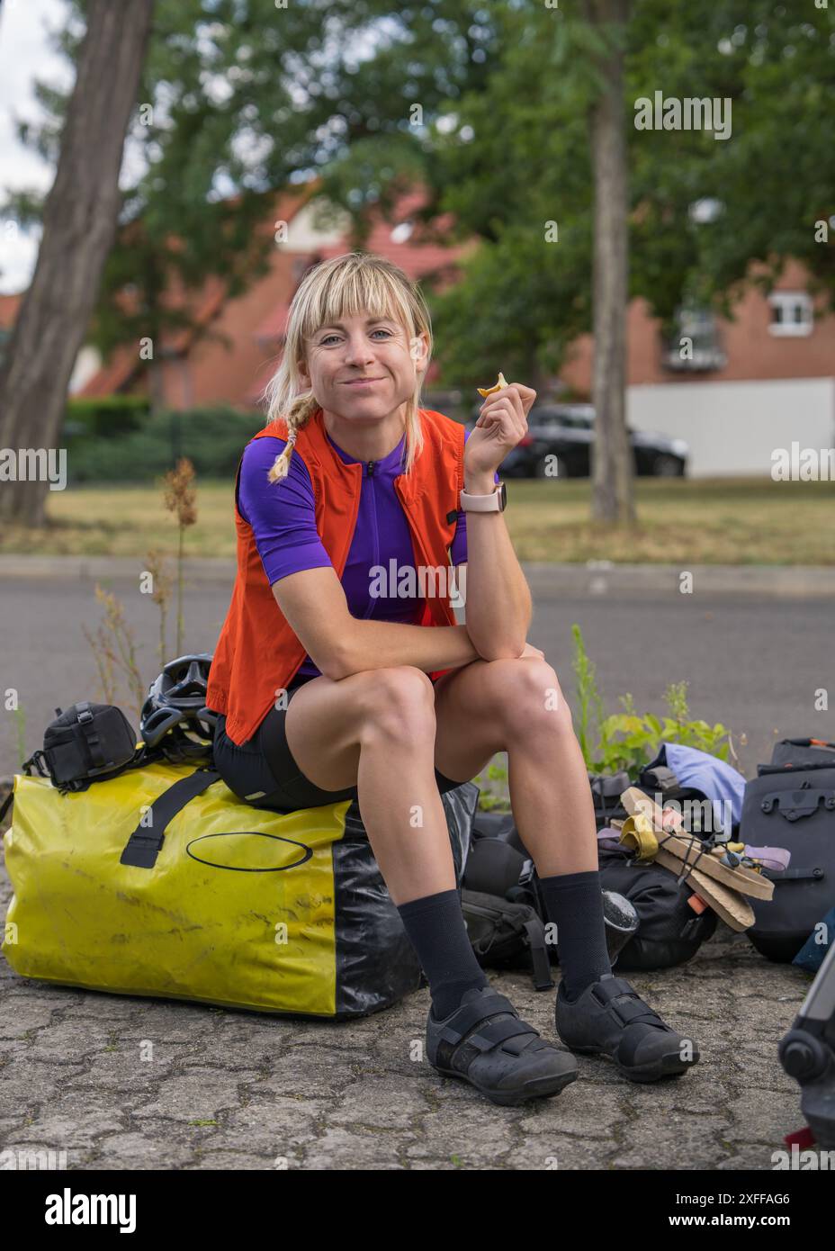 Die Radfahrerin hält an, sitzt auf ihren Fahrradtaschen und isst. Müdigkeit, mehrtägige Fahrradtour. Aktiver Lebensstil. Stockfoto