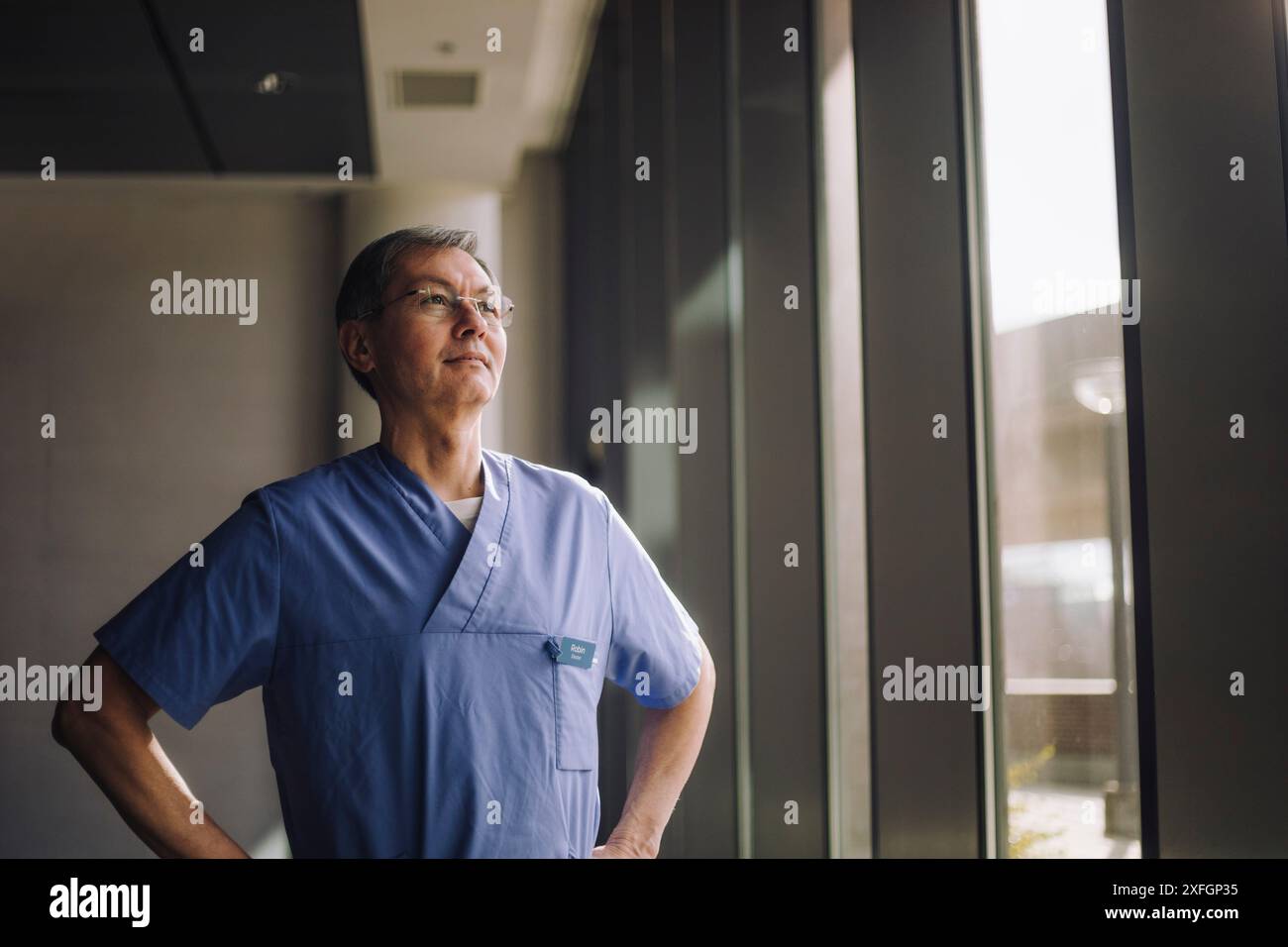 Selbstbewusster Senior männlicher Arzt stehend mit Armen akimbo nahe Fenster im Krankenhaus Stockfoto