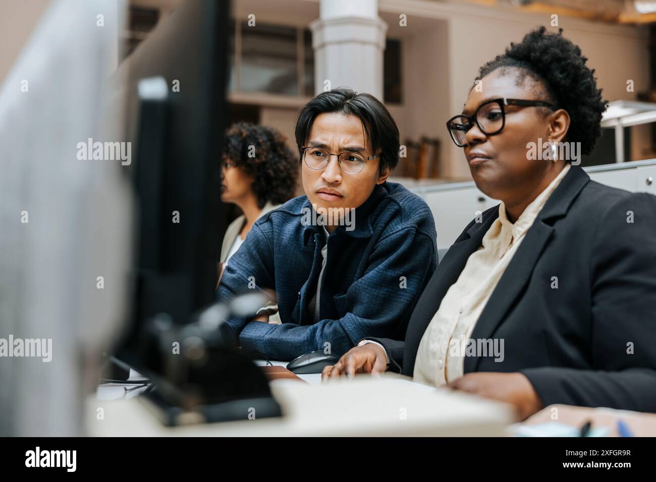 Ein konzentrierter Geschäftsmann sitzt mit einer Kollegin, während er im Büro den Computerbildschirm teilt Stockfoto