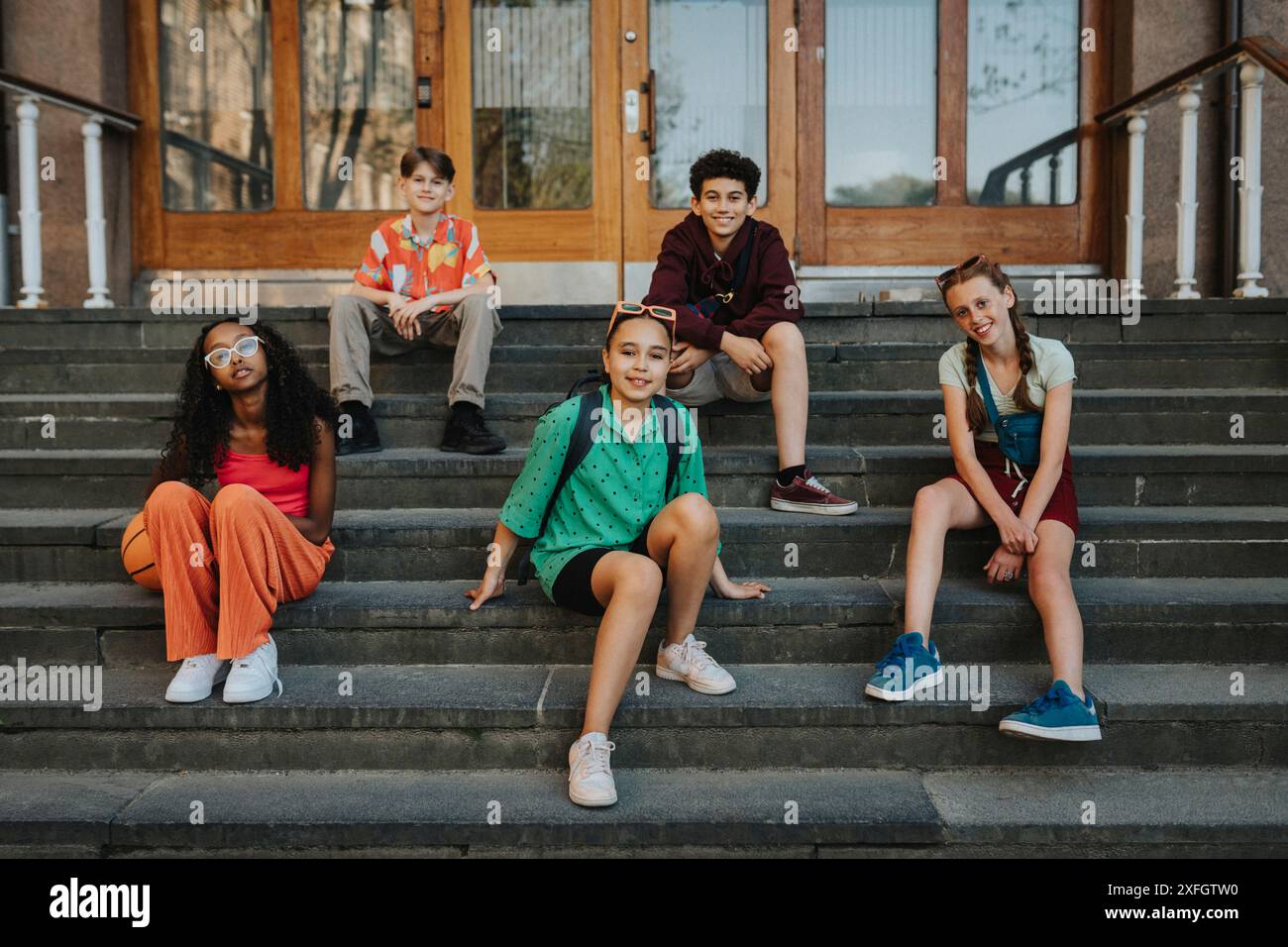 Glückliche Gruppe von Freunden, die auf der Treppe des Eingangs des Schulgebäudes sitzen Stockfoto