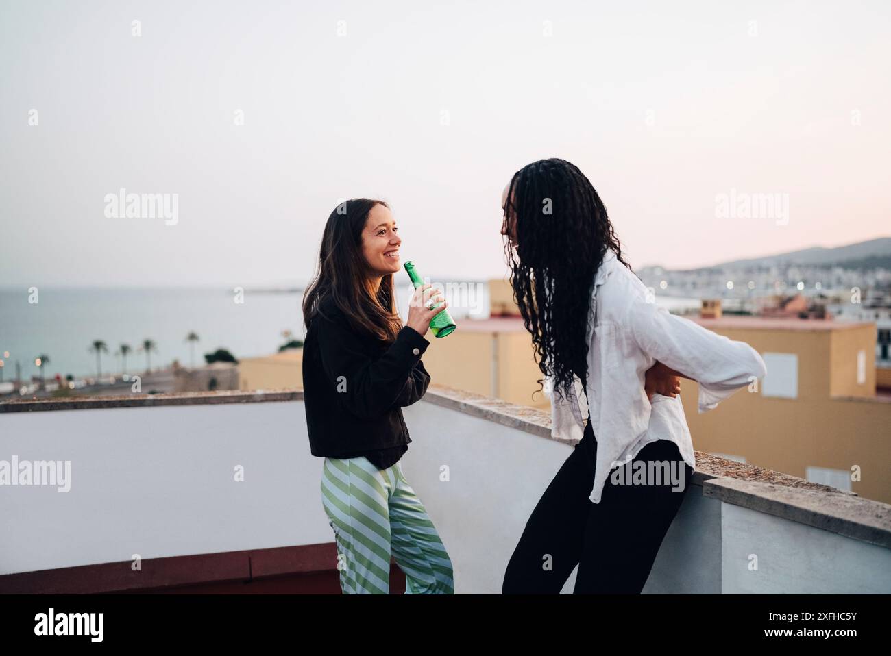 Lächelnde junge Frau trinkt Bier, während sie mit einer Freundin auf der Dachterrasse spricht Stockfoto