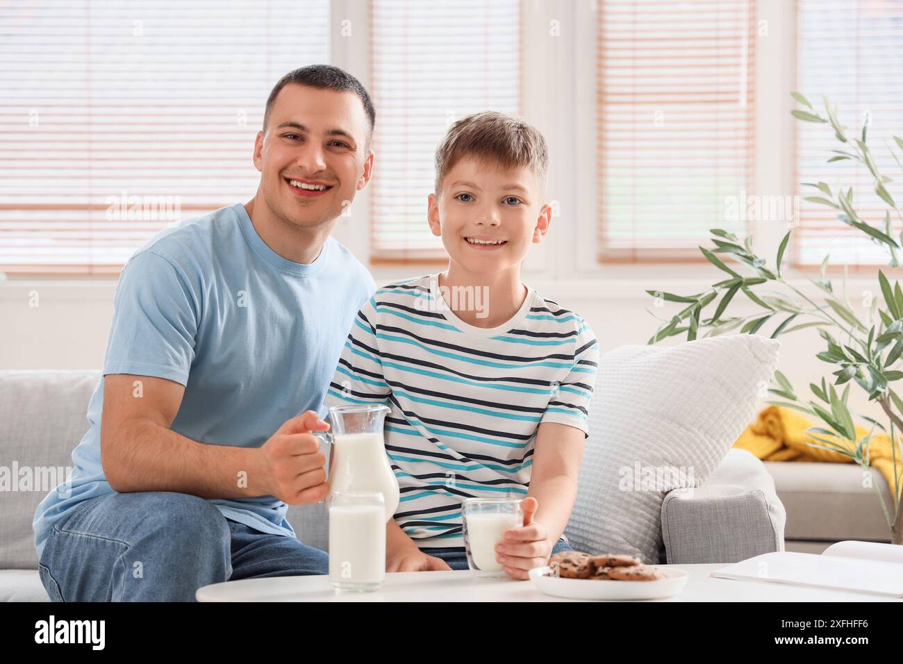 Glücklicher Vater mit einem Krug Milch und sein kleiner Sohn sitzt zu Hause auf dem Sofa Stockfoto