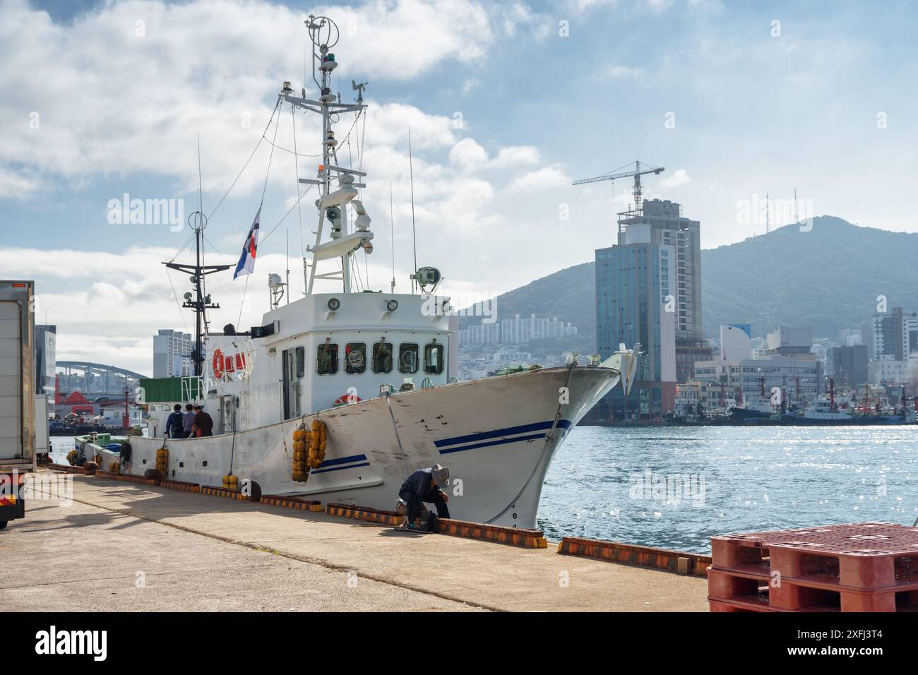 Geparktes weißes Schiff im Hafen von Busan in Südkorea. Busan City ist auf dem Hintergrund der Berge zu sehen. Stockfoto