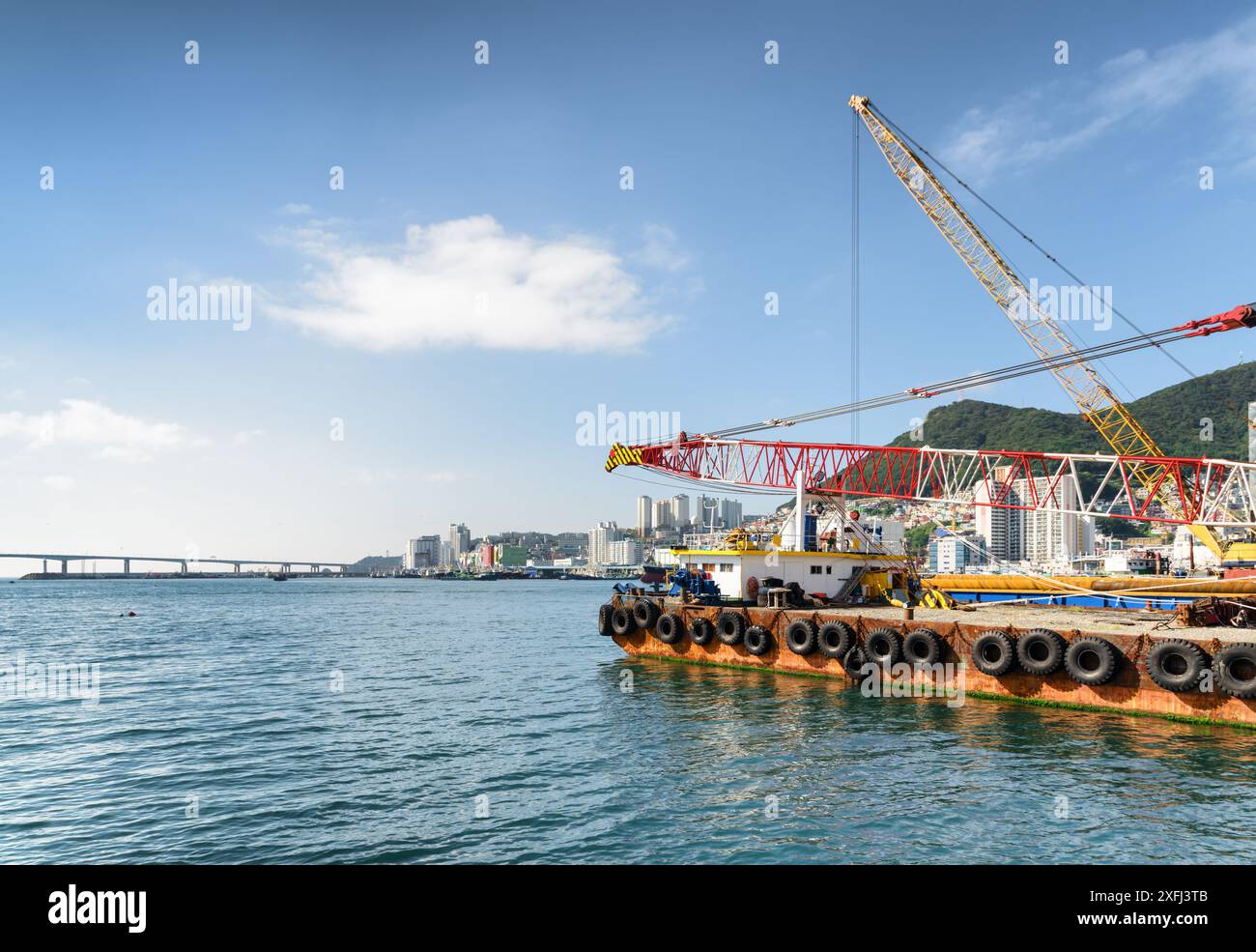 Kranschiff auf blauem Himmel im Hafen von Busan in Südkorea. Busan City und Namhangdaegyo Bridge sind im Hintergrund zu sehen. Stockfoto