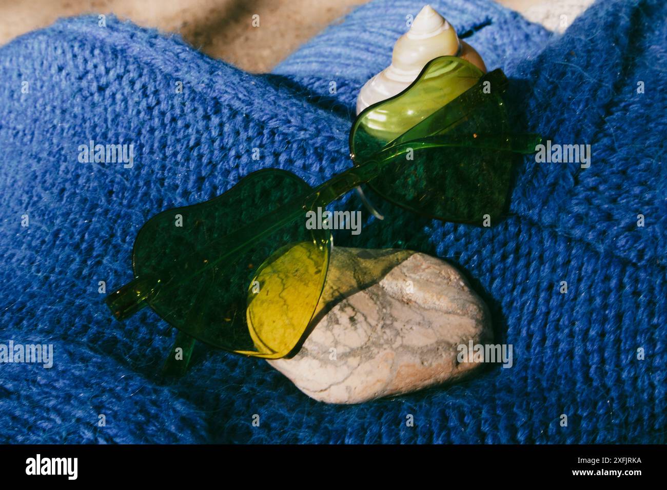 Grüne Sonnenbrille in Herzform, Muschel auf blauem Textil. Urlaub am Meer, Meer im Sommer. Entspannen im Sommer. Weibliches Zubehör. Sonniger Tag. Stockfoto