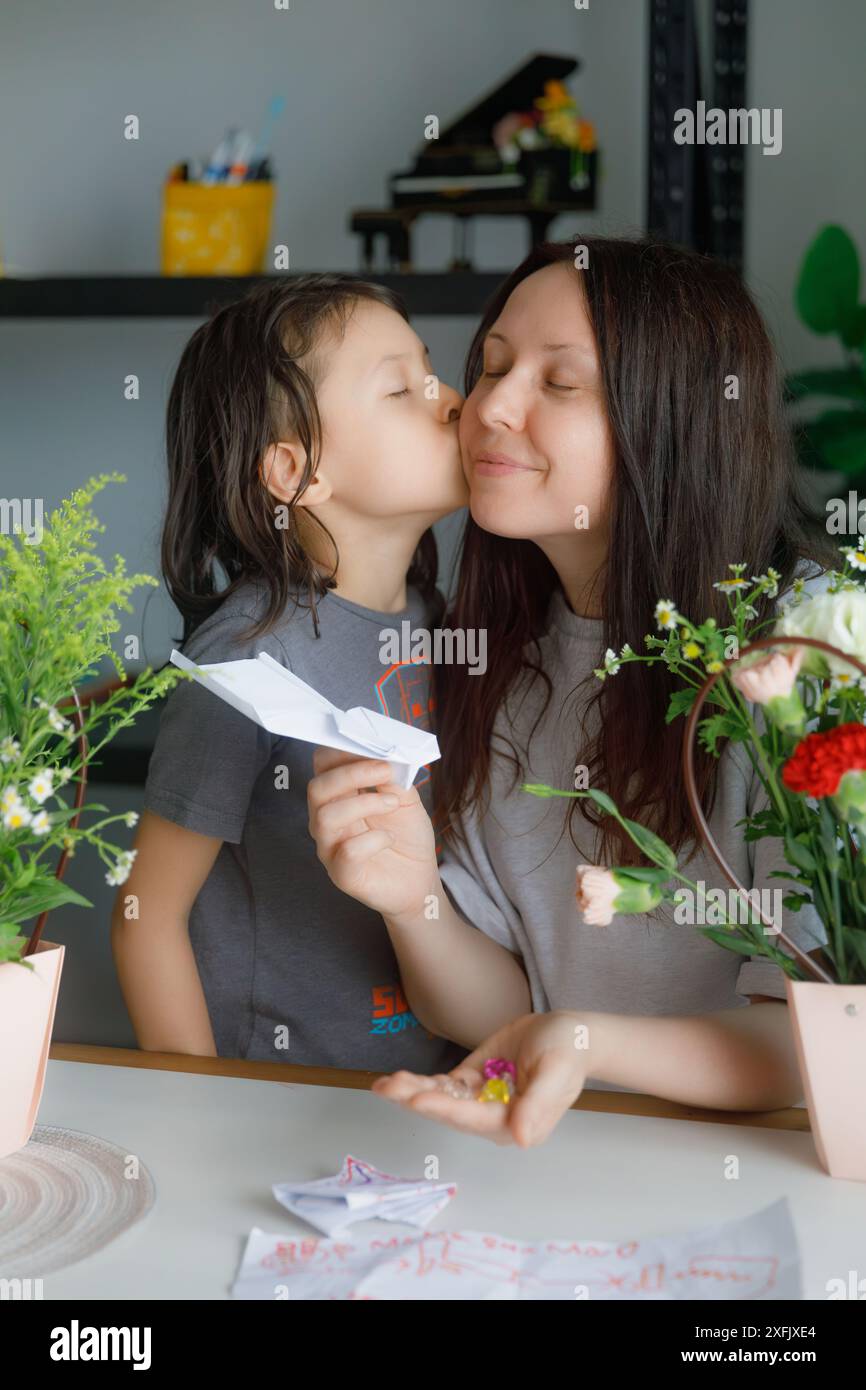 Der Sohn küsst seine Mutter. Die junge Frau lächelt glücklich, während sie das Muttertagsgeschenk zeigt, das ihr Sohn ihr gegeben hat. Stockfoto