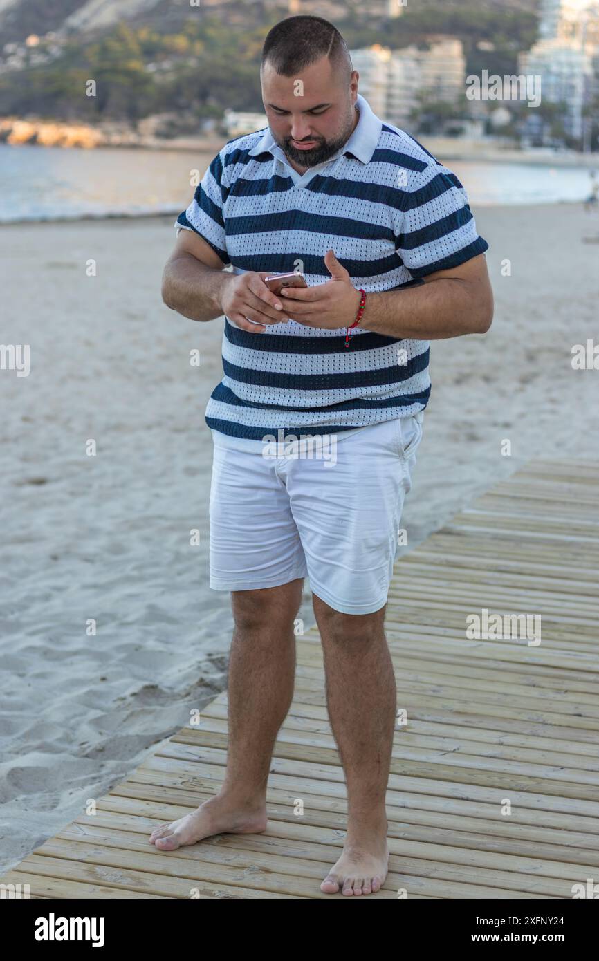 Dieses Bild zeigt einen Mann, der an einem Strand steht und in sein Smartphone vertieft ist. Seine lässige Kleidung lässt einen gemütlichen Tag vermuten. Stockfoto