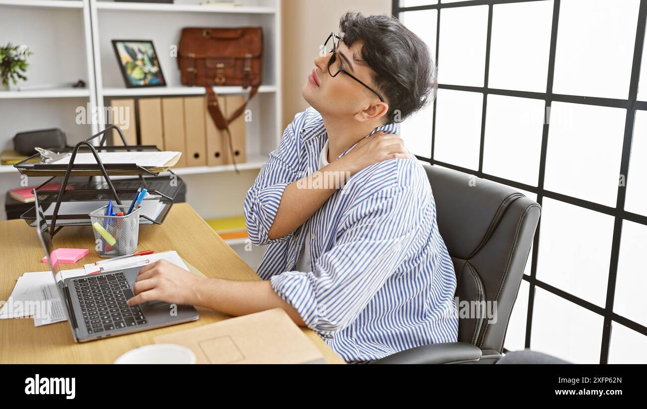 Junger Mann in gestreiftem Hemd, der Nackenschmerzen spürt, während er in einem modernen Büro am Laptop arbeitet. Stockfoto