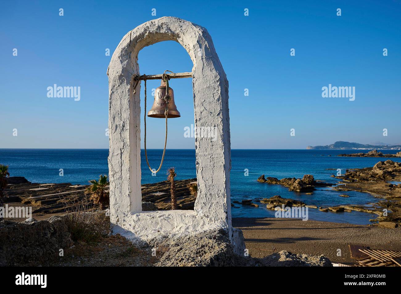 Weißer Glockenrahmen mit Glocke vor einer felsigen Küste und blauem Himmel, Palmen im Hintergrund, Felsenkirche Agios Nikolaos, Oasis Beach, Kallithea Stockfoto