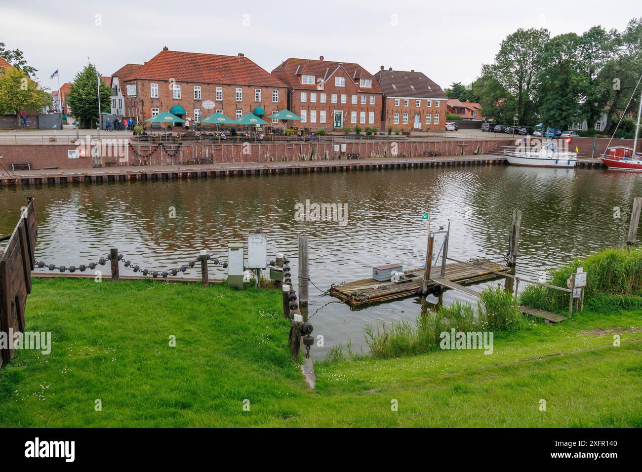 Uferufer mit alten Backsteinbauten und einer Holzplattform im Wasser, einige Boote am Ufer, wangerland, niedersachsen, deutschland Stockfoto