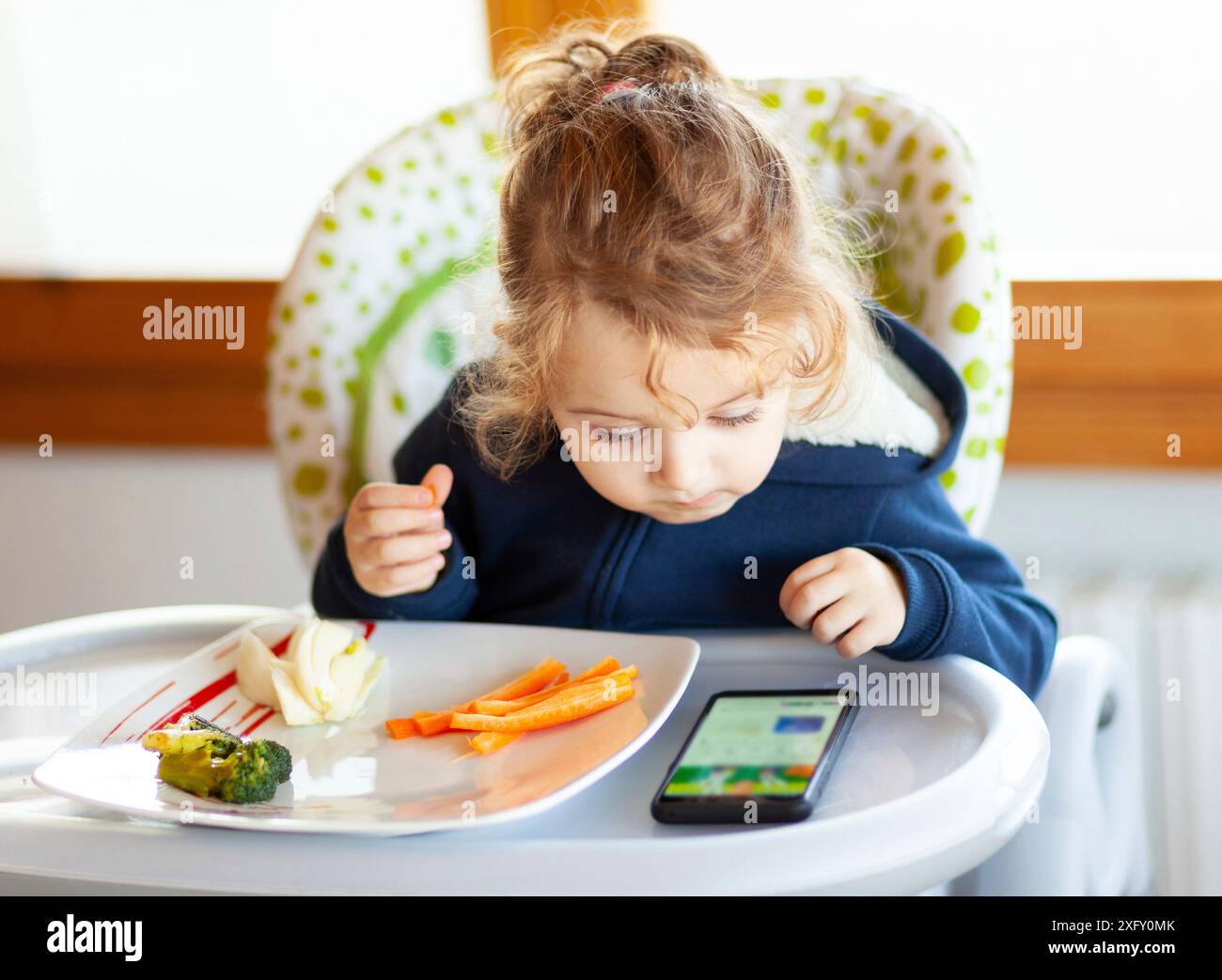 Kleinkind isst im Hochstuhl, während es Filme auf dem Handy sieht. Viele Kinder wollen nicht essen, wenn sie nicht auf das mobile Gerät oder Fernsehen. Stockfoto