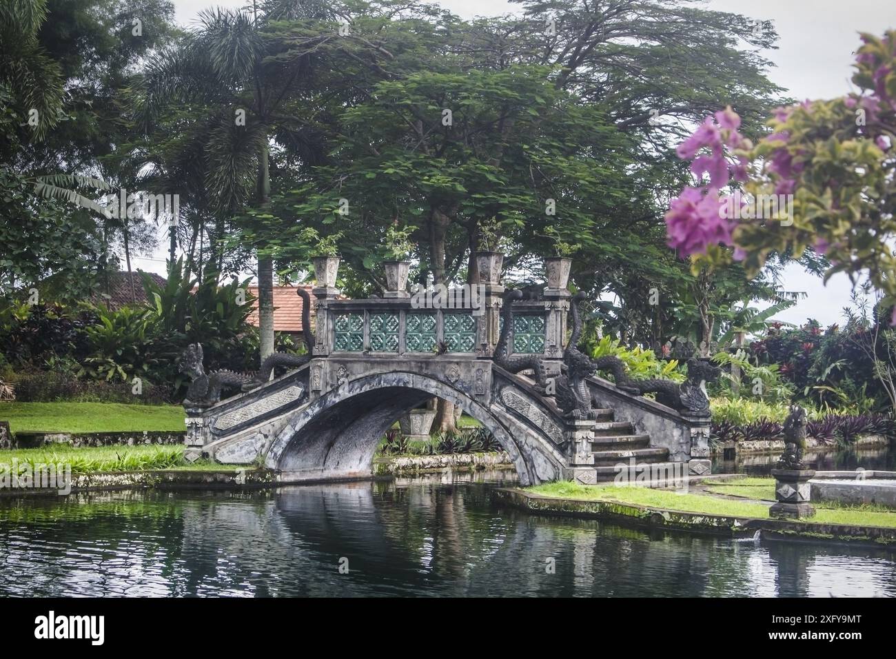 Steinbrücke über den Fluss im Wasserpalast Tirtagangga auf Bali. Steinstatuen, üppige grüne Vegetation und Wasserfontänen. Stockfoto