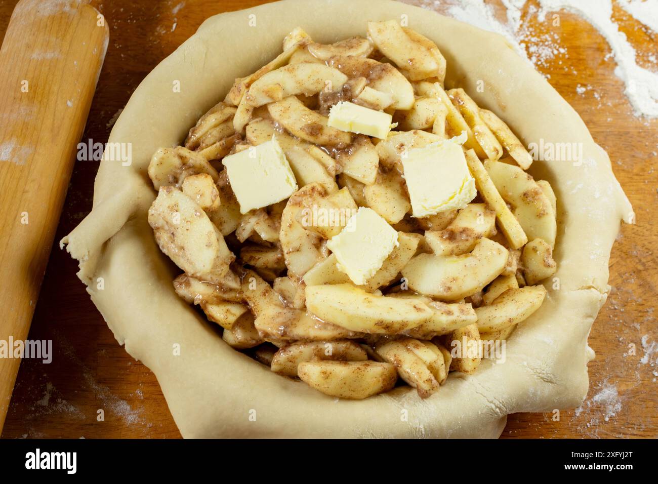 Torte machen. Gerollter Teig über eine Kuchenschale gelegt und mit Apfelscheiben gefüllt, die mit Butter gefüllt sind. Ansicht von oben nach unten. Rollstift im Hintergrund. Stockfoto