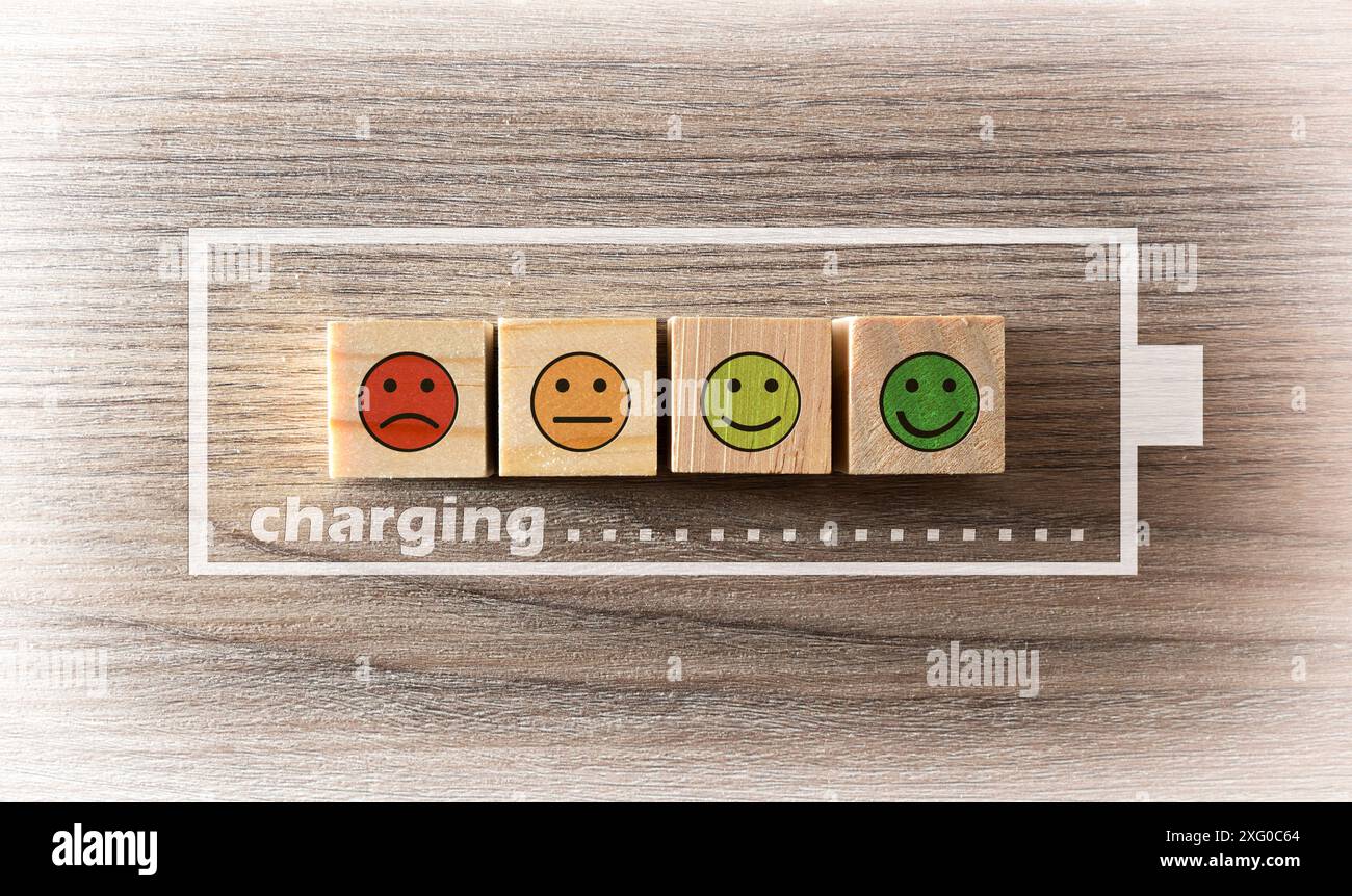 Konzept der Aufladung der Vitalenergie mit Holzblöcken mit Emoticons mit Ausdrücken auf Holztisch und Batteriesymbolen und Ladesymbolen. Stockfoto
