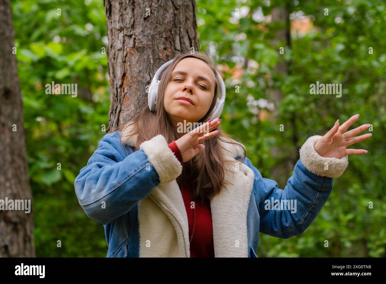 Eine junge Frau lehnt sich an einen Baum, trägt Kopfhörer und genießt Musik im Wald an einem sonnigen Nachmittag Stockfoto