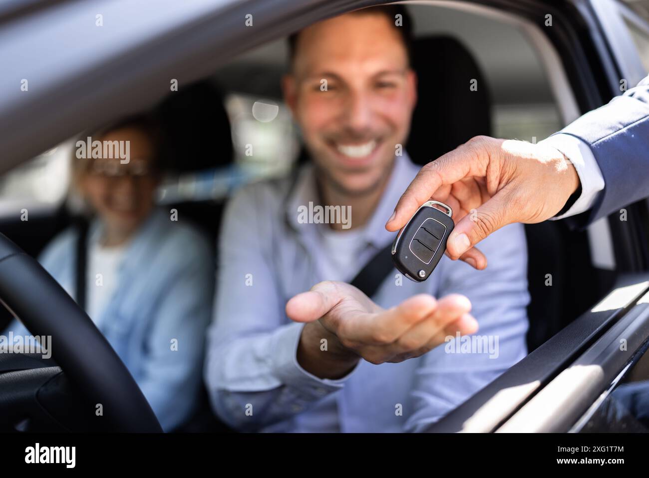 Ein lächelnder hispanischer Verkäufer übergibt erfolgreich während der Geschäftszeiten Schlüssel an Neuwagenbesitzer im örtlichen Händlerbetrieb. Stockfoto