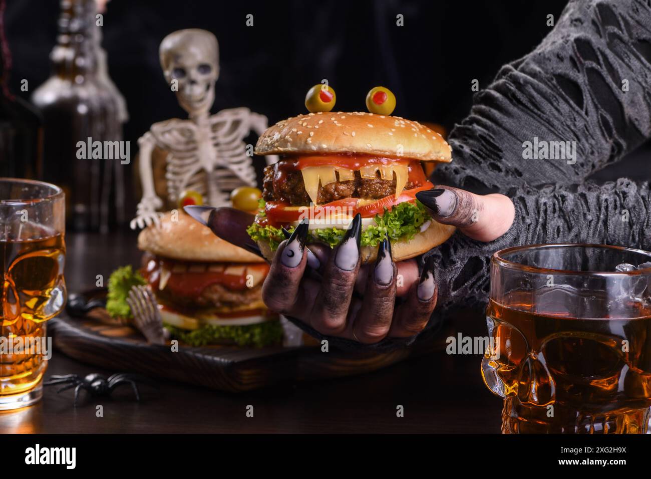 Die Hexenhände halten einen Monster Burger in ihren Händen auf einem sitzenden Skelett. Die perfekte Vorspeise für die Halloween Party Stockfoto