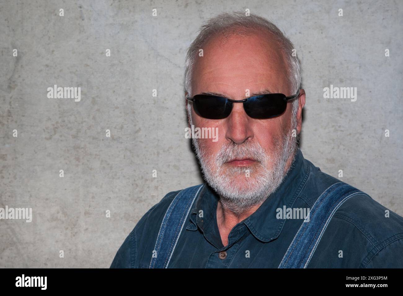 Ein älterer Mann mit grauem Haar, dunkler Sonnenbrille und Vollbart steht vor einer Betonmauer. Sein ausdrucksloser Blick erscheint bedrohlich. Die Co Stockfoto