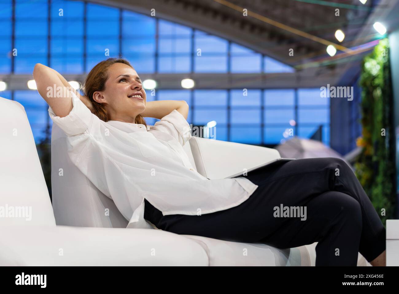 Junge Erwachsene Geschäftsfrau mit Händen hinter dem Kopf, die sich auf dem Sofa in der Business Lounge des Flughafens oder im Büro entspannen. Stockfoto
