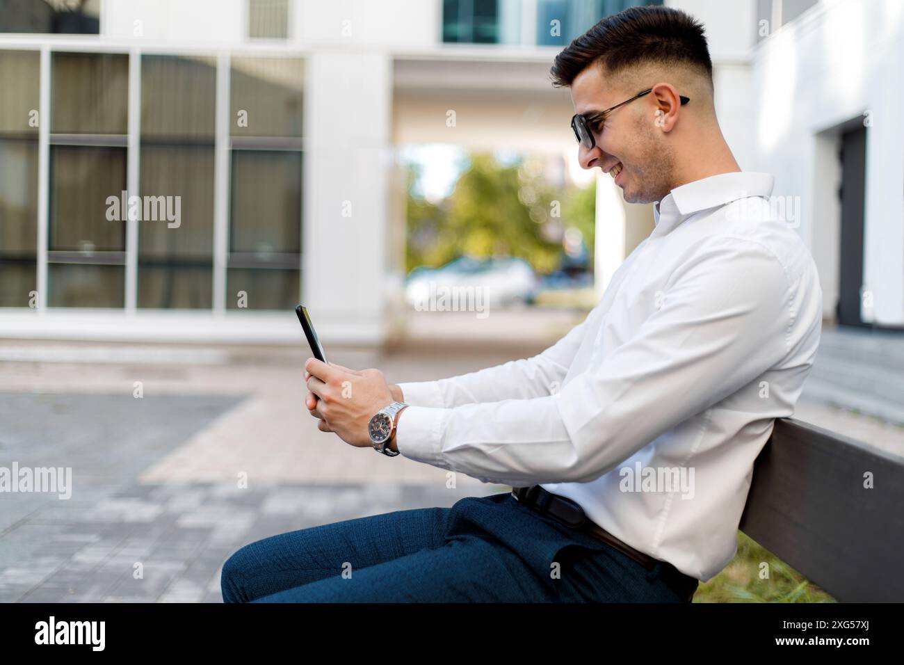 Ein Mann sitzt auf einer Bank, vertieft in seinen Handy-Bildschirm. Er scheint auf das Gerät konzentriert zu sein, möglicherweise liest er Nachrichten oder sucht nach. Stockfoto