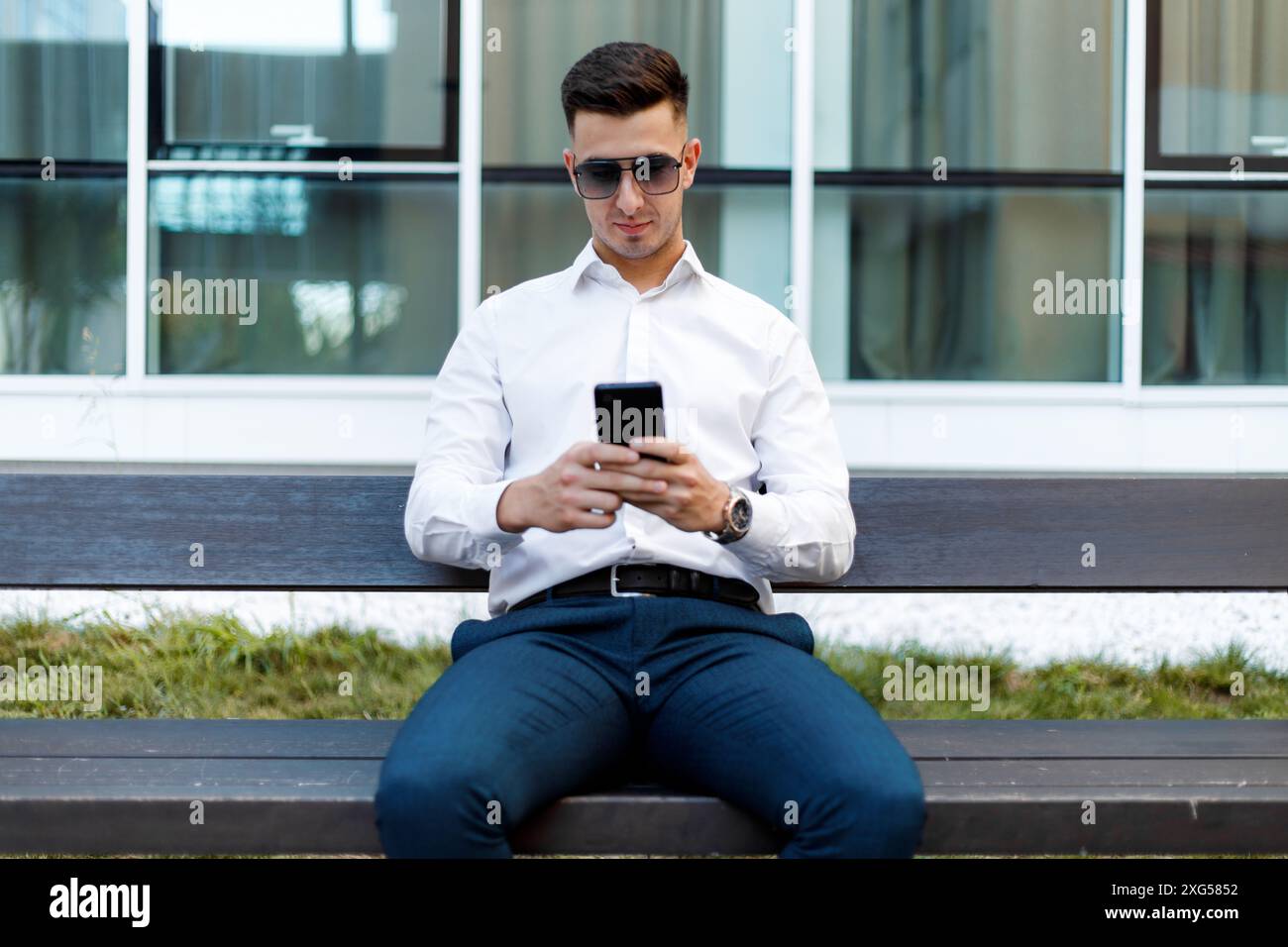 Ein Mann sitzt auf einer Bank, vertieft in sein Handy. Er scheint auf dem Bildschirm konzentriert zu sein, wobei das Gerät in beiden Händen gehalten wird. Stockfoto