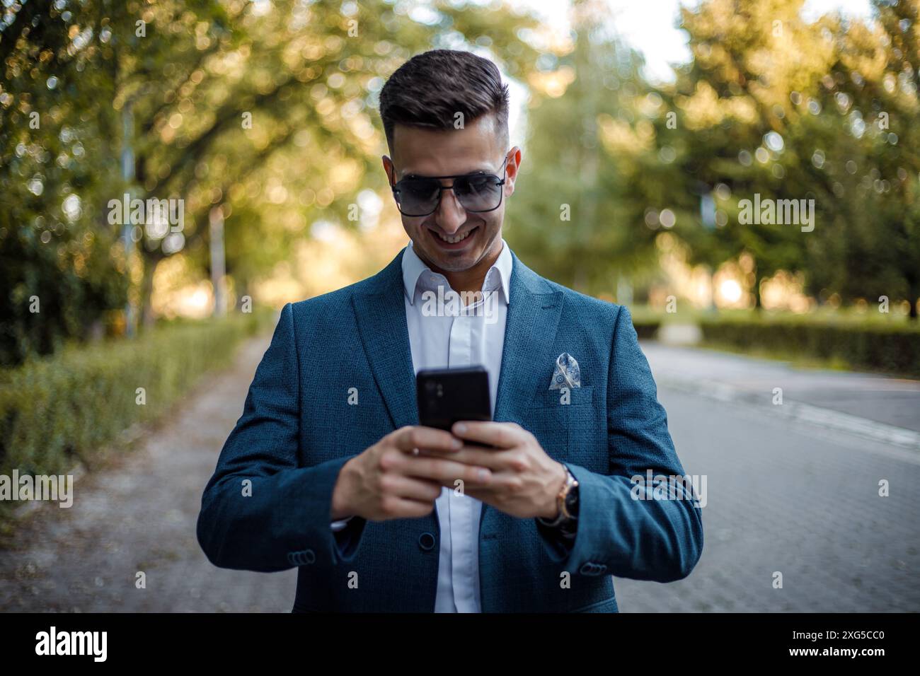 Ein Mann, der einen Anzug trägt und eine Sonnenbrille trägt, konzentriert sich auf seinen Handy-Bildschirm, scrollt und tippt aufmerksam. Er steht in einer geschäftigen städtischen Umgebung. Stockfoto